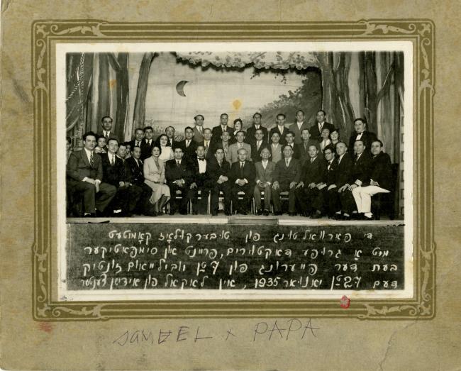 A group of formally dressed adults pose on a stage with a theatrical set in the background. Photo captioned in Yiddish.