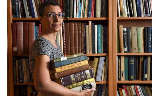 Person stands in front of shelves of books holding a stack of books and looking at the camera