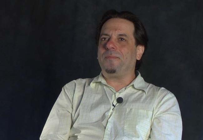 Man in white button up shirt sits in front of dark background and looks at camera