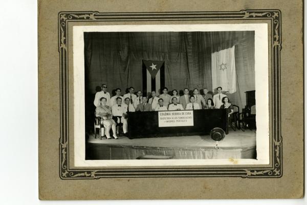 A photo of adults seated at a table labeled Colonia Hebrea de Cuba, Protectora De Los Tuberculosos y Enfermos Mentales with a Cuban flag and a flag with a Star of David behind them.