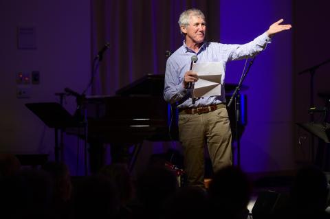 Man wearing blue shirt and khakis stands onstage with purple background lighting.