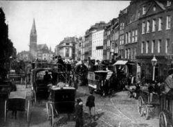 Black and white image of a Whitechapel street, crowded with people and carriages.