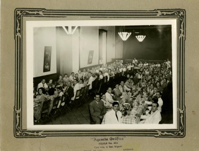 Adults and children sit at two long tables in a social hall. English, hand-written caption on the left reads " Banquet for Pasiencier 1942."