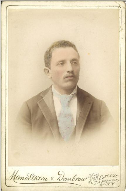 Colorized portrait of a young man with a moustache, wearing a blue necktie and black suit. Text reads: Mandelkern and Dombrow, 183 Essex St, cor. Houston St., N.Y.
