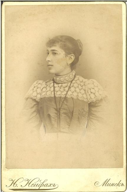 Old sepia-toned photograph of a your woman wearing a dress with lace on the shoulders. Her hair is in a bun. Text reads; N. Neyfakh, Minsk, in Cyrillic.