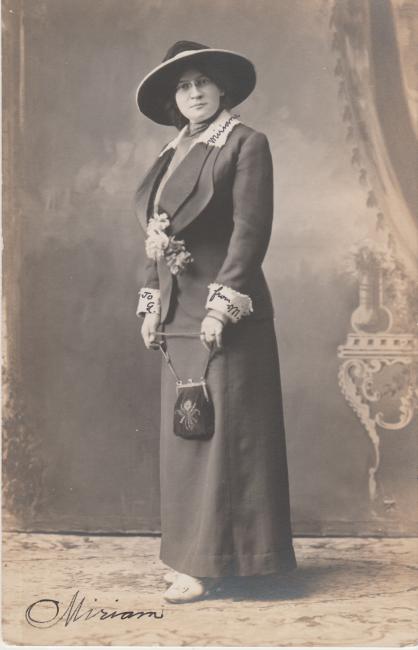 Black and white studio photograph of Miriam Karpilove, wearing a black dress with lace trim and a black hat, holding a purse