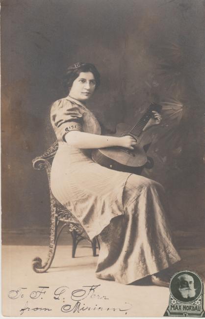 Black and white studio photograph of Miriam Karpilove, wearing a white dress, sitting in a chair playing the guitar.