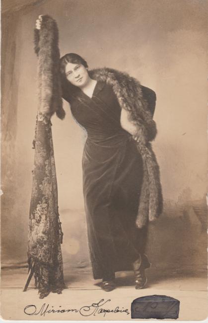 Sepia-toned portrait of Miriam Karpilove, standing wearing a black dress, holding a fur.