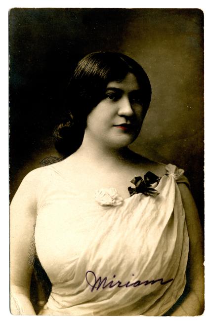 Young Miriam Karpilove sitting for a black and white portrait, with her hair in a bun, wearing a white toga-style dress. The image is signed "Miriam".