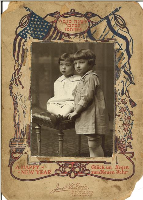 Rosh-Hashana greeting card with a photo of two young children, one seated and one standing, bordered by American and Zionist flags and the Statue of Liberty. Text reads: leshana tova tikoseyvu vetakhateymu, a happy new year, Gluck un Segen zum neuen Jahr, Jacob E. Dwin, 1697 Pitkin Ave., cor. Rockaway Ave., Brooklyn, N.Y.
