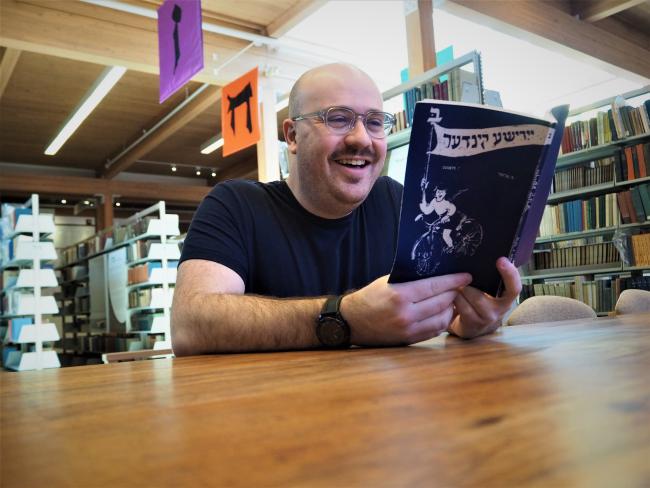 Caleb, bald white man wearing a blue t-shirt, reads a Yiddish learning book.