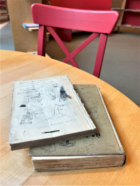 Two graffitied books sit on a wooden table in front of a red chair.