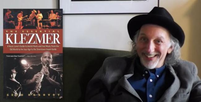 Seth Rogovoy sits on a couch in a grey coat with his book The Essential Klezmer