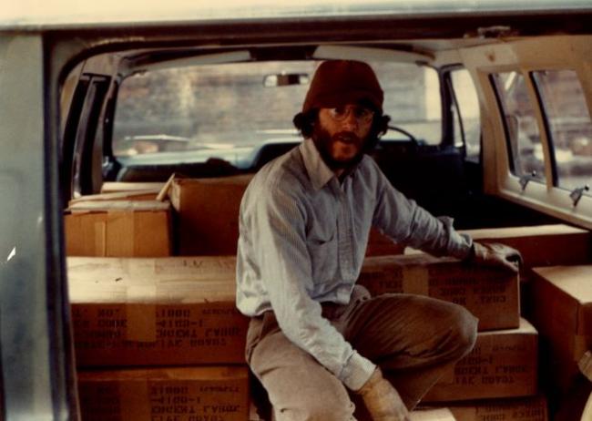 A man with glasses sits on a stack of boxes in the trunk of a car