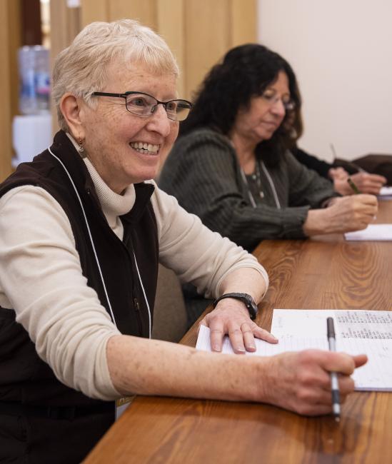 Woman with white hair and glasses wearing black sweater vest smiles sitting at desk. Dark haired woman sits in background of image.