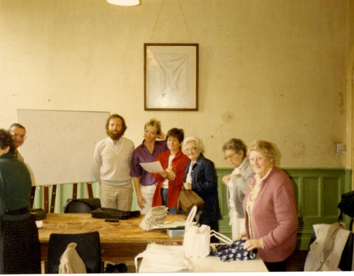 Eight adult students stand in a Yiddish classroom with a table and whiteboard.