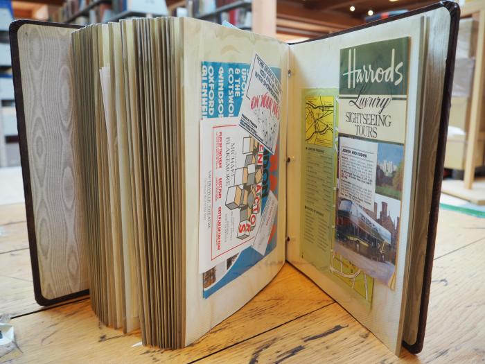 A picture of the large scrapbook standing up on a table, open to a page with brochures from sites in England such as Harrods.