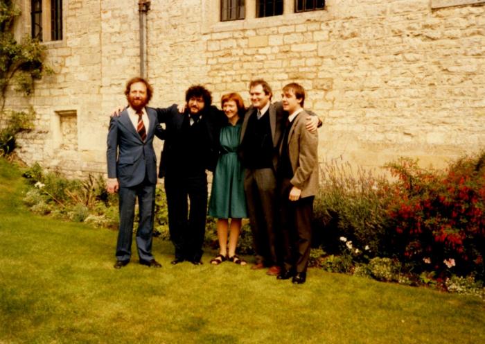 Picture of four men and one woman in the center, arms wrapped around each other, standing on the lawn in front of a white stone building.