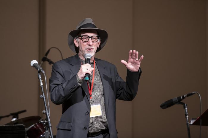 Man wearing glasses and fedora hat holds microphone and speaks to audience