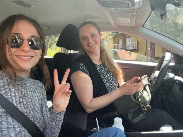Two women sit in a car, one makes a peace sign and both smile at camera