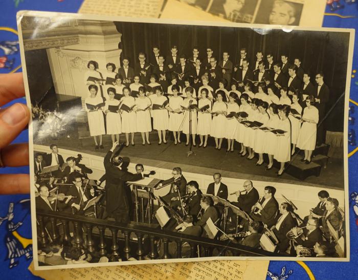Jewish choir with women wearing white dresses and men wearing dark suits. A conductor and band plays in front of them.