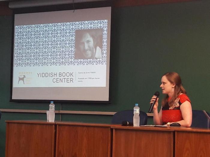 Christa Whitney wearing a red shirt holds a microphone while sitting behind a desk and presents a slideshow on the Yiddish Book Center.