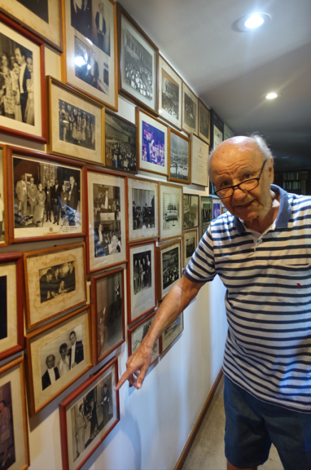 Man wearing glasses and blue and white striped polo shirt shows photographs hanging on a wall.