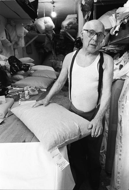 Man wearing glasses, white undershirt, and suspenders poses with feather pillows in workshop