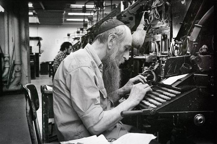 Man wearing kippah with long beard works at a typesetting machine, black and white photo.