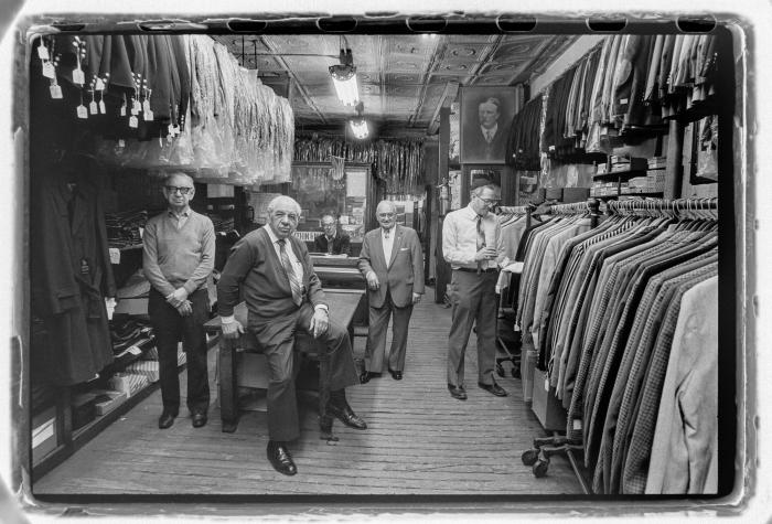 Five men wearing suits and ties pose in a suit warehouse room, black and white photo.