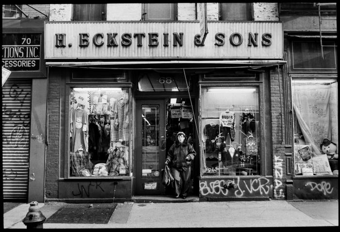 Woman wearing fur-trimmed coat walks out of NY store H. Eckstein & Sons, black and white photo.