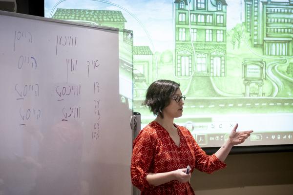 Woman with short hair gesturing in front of a projector and whiteboard with Yiddish writing on it