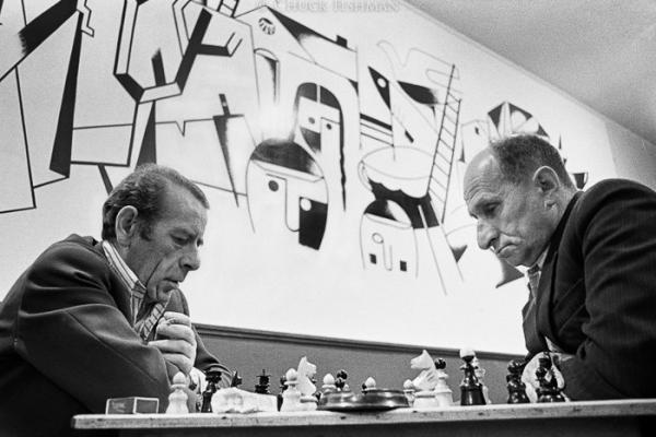 Two men play chess in black and white photo with mural behind. 