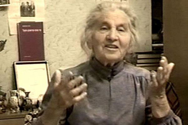 Shira Gorshman speaking, an older woman with grey hair and a purple blouse. Her book is mounted in the background.