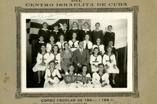 Children in uniforms sit, with two adults in the center. Two children at the front hold a banner reading "5th grade" in Spanish