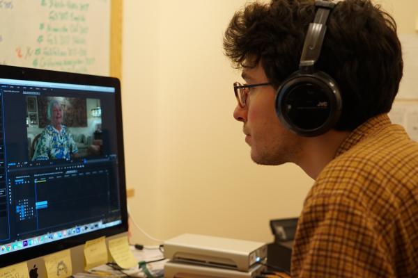 Person with brown hair, glasses, and headphones looking at computer screen with interview playing