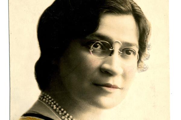 Colorized portrait of Miriam Karpilove wearing glasses and a pearl necklace. Cropped around her face.