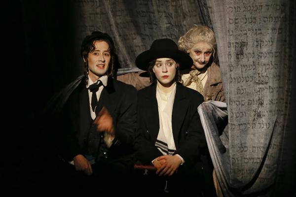 Three actors in costume posed with a grey curtain with Yiddish text