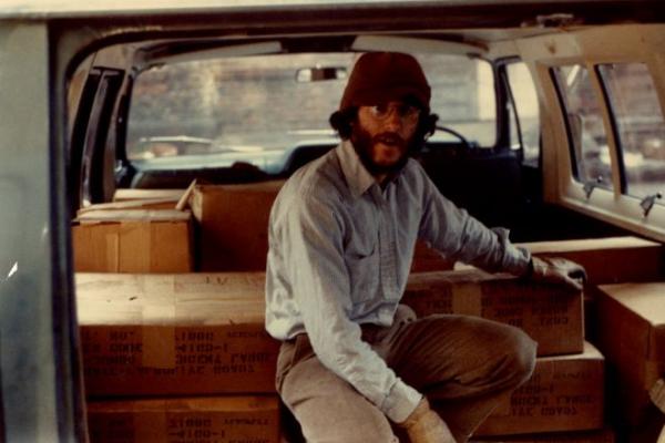 A man with glasses sits on a stack of boxes in the trunk of a car